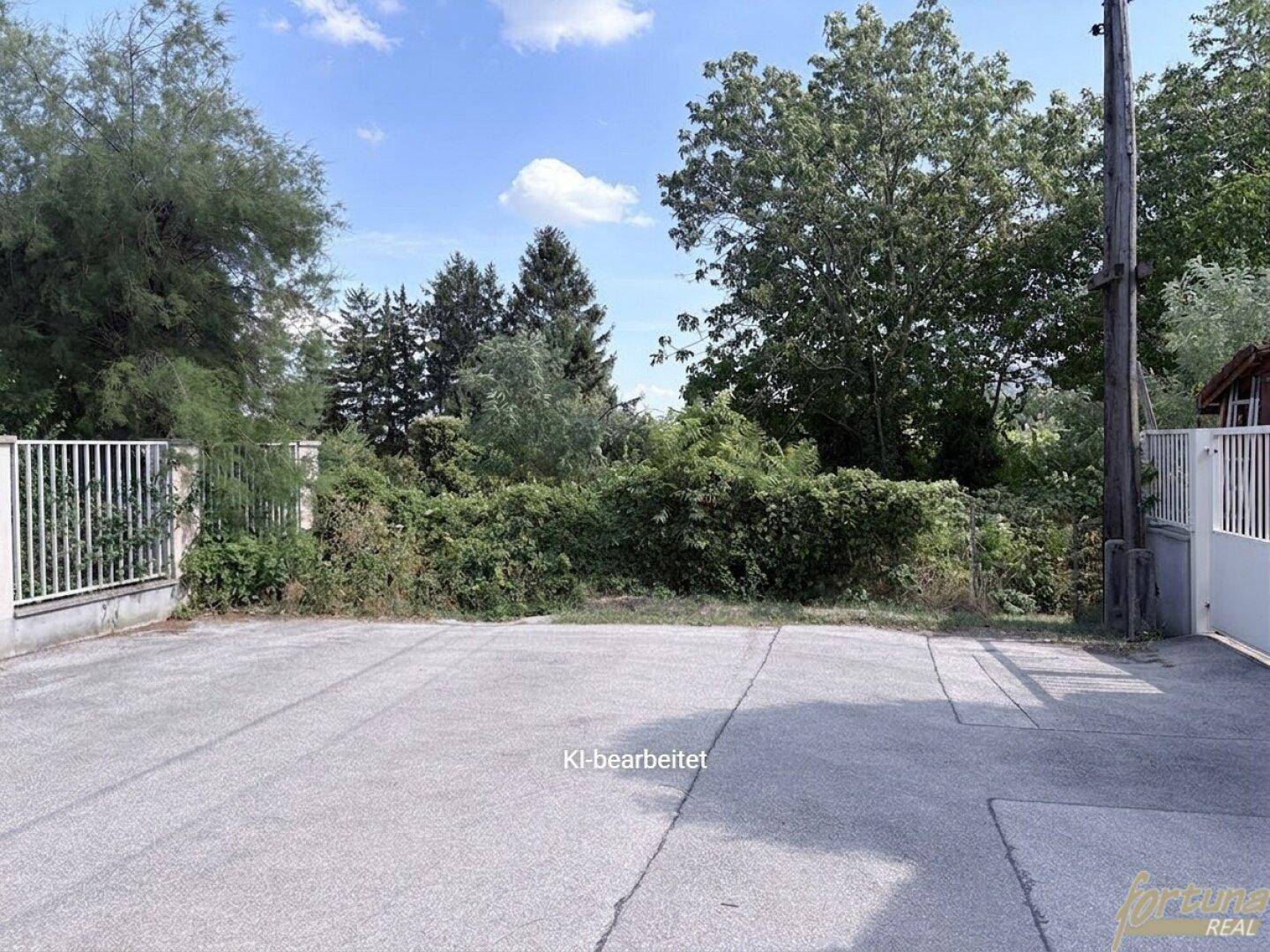 Große, gepflasterte Fläche, umgeben von Zäunen und Bäumen, möglicherweise ein Parkplatz.