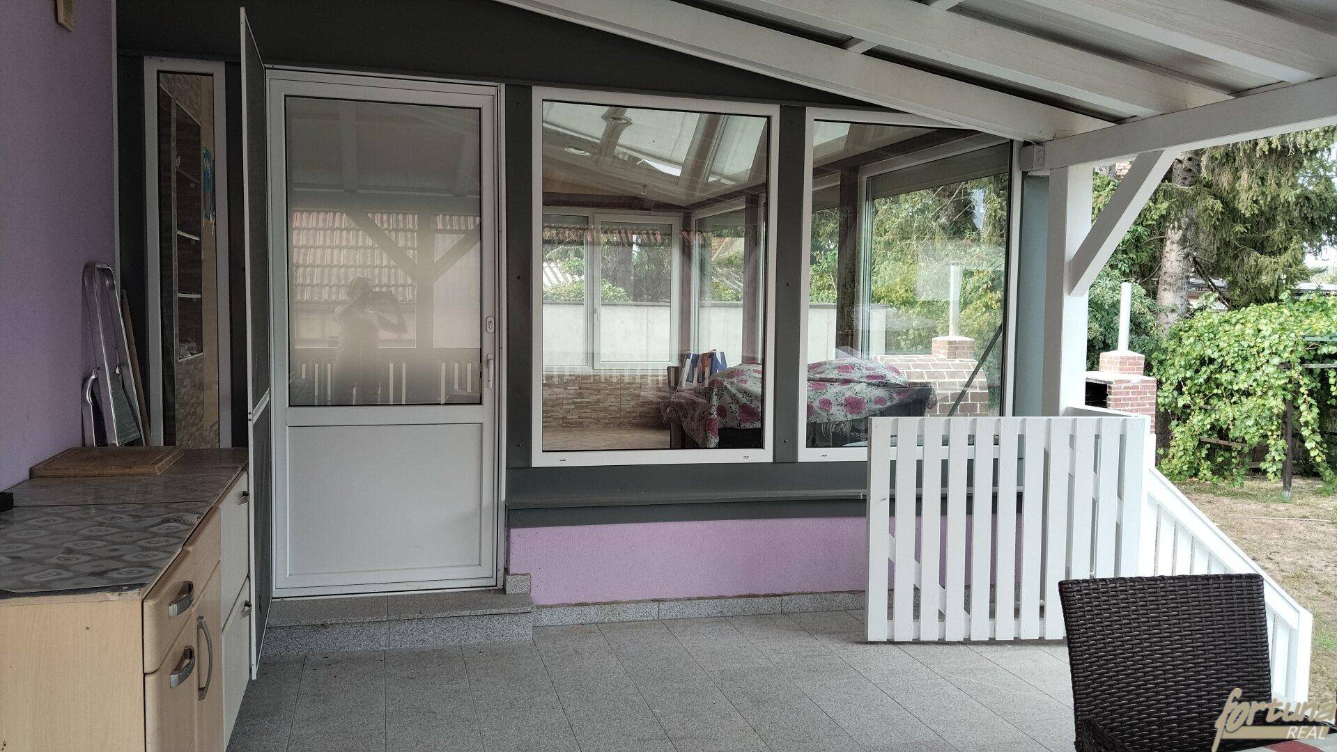 Überdachte Terrasse mit Zugang zum Wintergarten und einer kleinen Außenküchenzeile, umgeben von Natur.