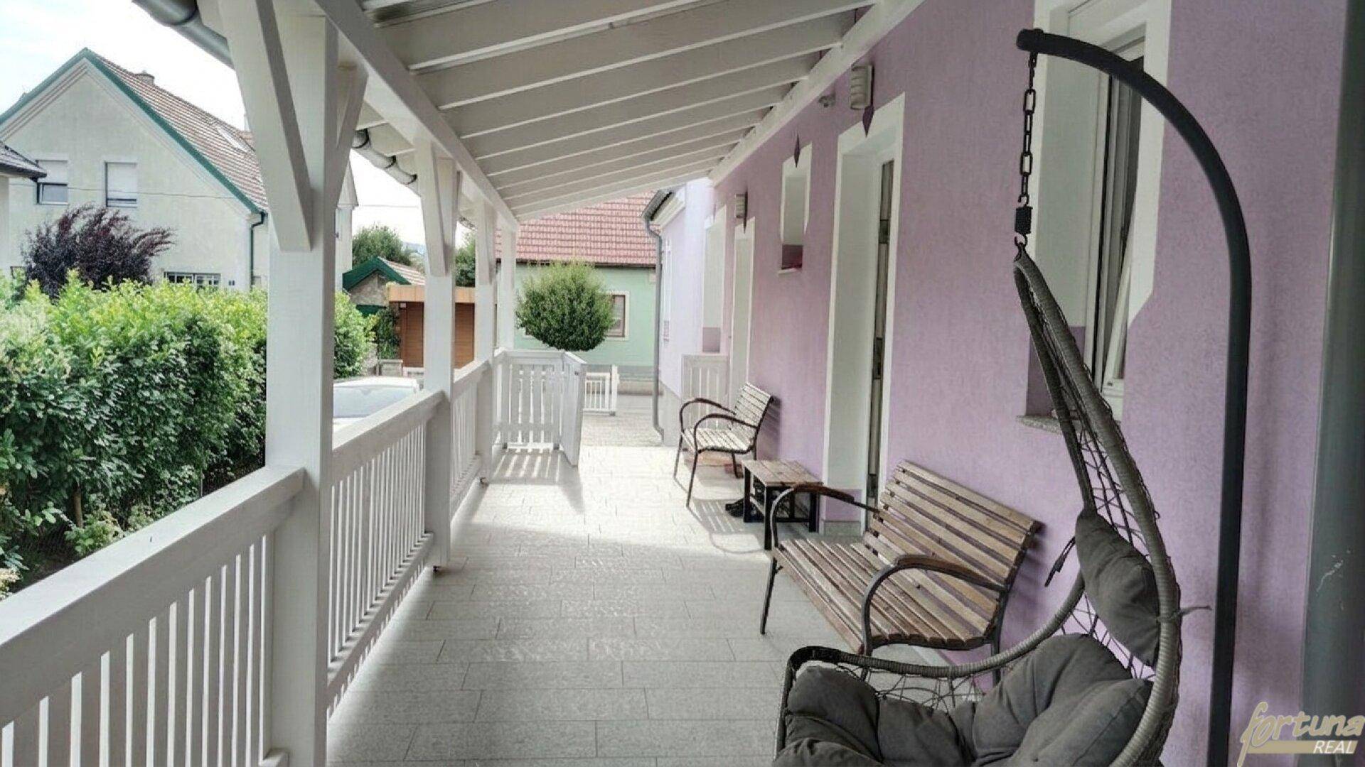 Langer überdachter Balkon mit Sitzbänken und Hängesessel, bietet einen entspannten Außenbereich entlang der Fassade.