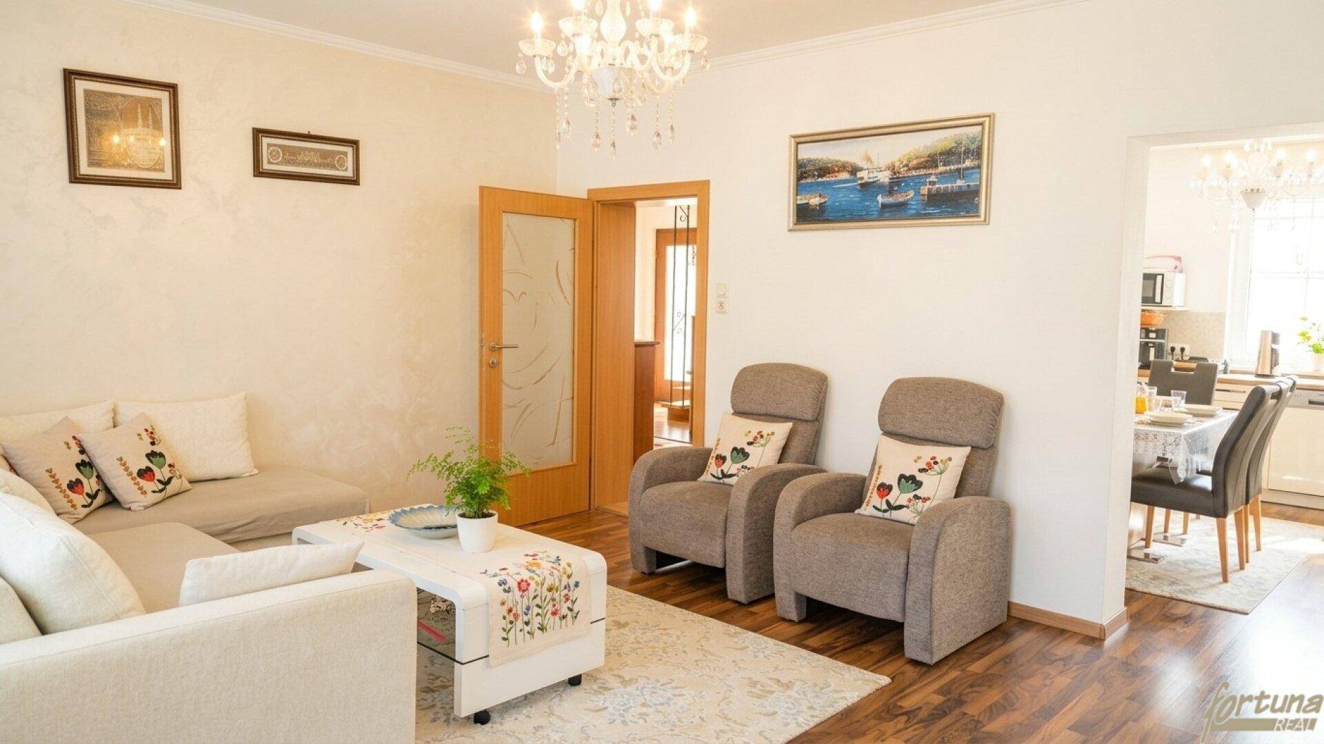 Gemütliches Wohnzimmer mit Sofa, Sesseln und Kronleuchter, offen zum Essbereich und zur Küche.