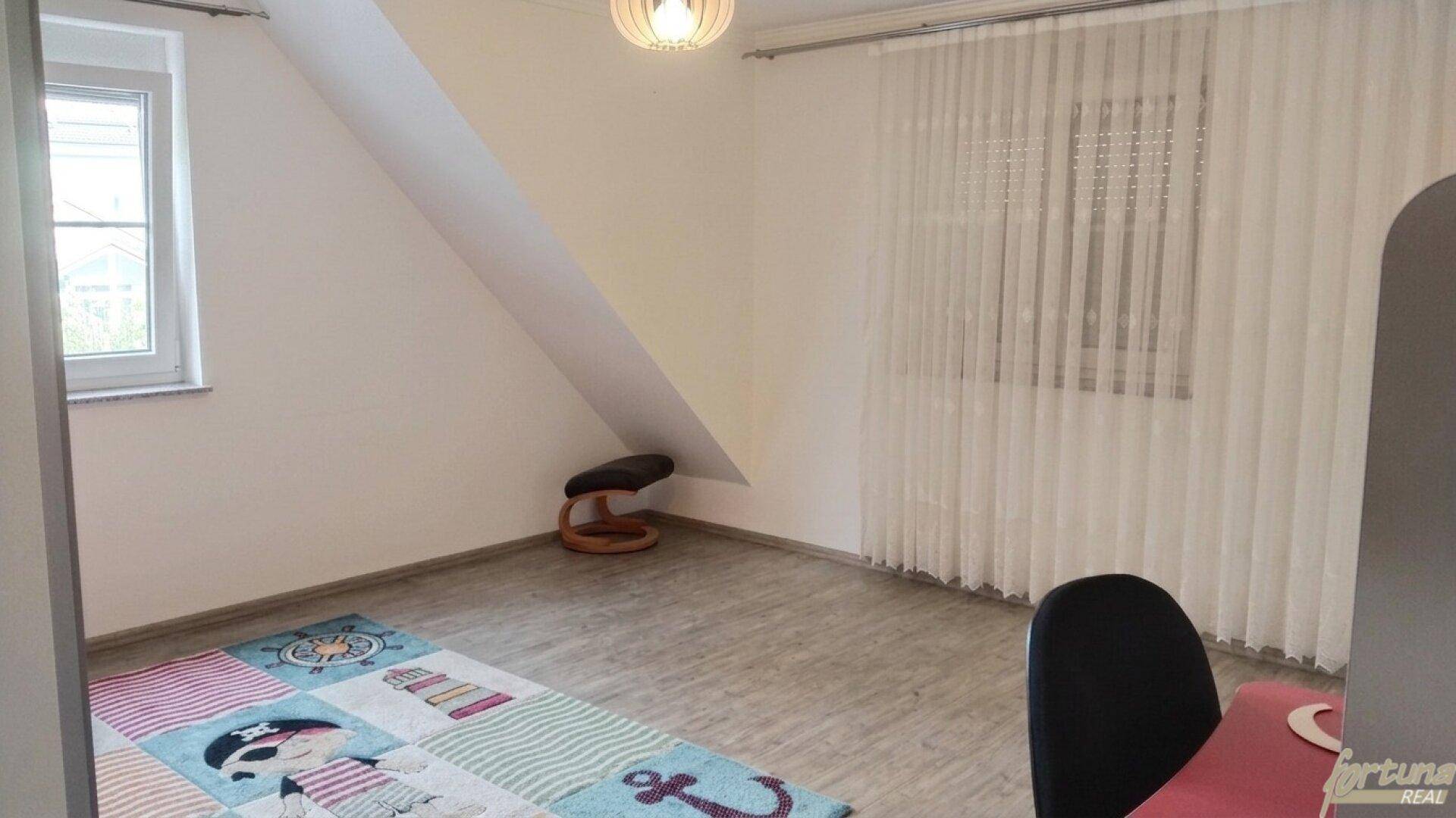Helles Zimmer im Dachgeschoss mit schräger Decke, Laminatboden und einem bunten Teppich.