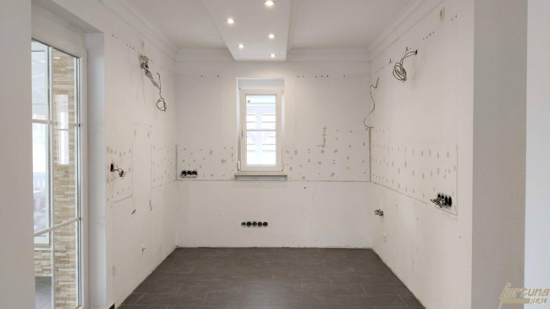 Leere Küche im Rohbau mit dunklem Fliesenboden und vorbereiteten Anschlüssen für die Kücheninstallation.
