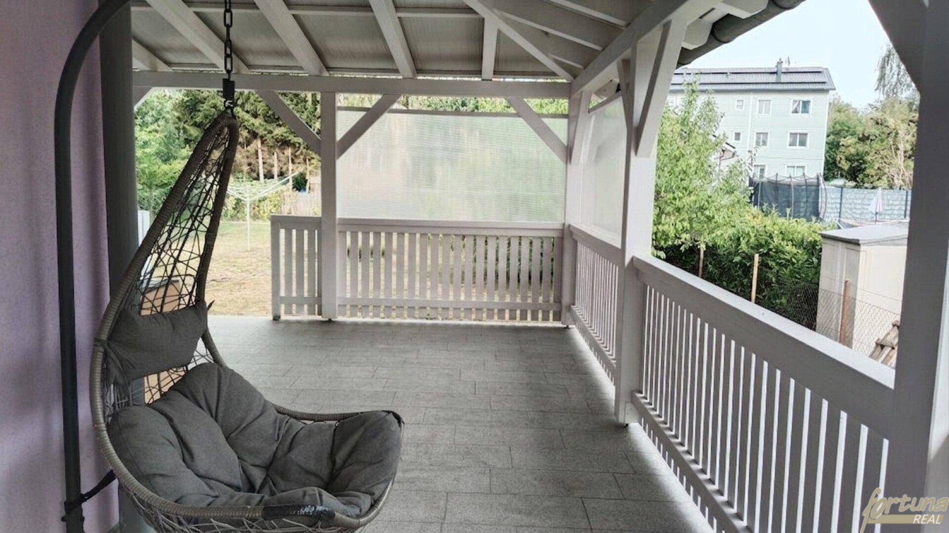 Überdachte Terrasse mit Hängesessel und Fliesenboden, bietet einen gemütlichen Außenbereich mit Blick ins Grüne.