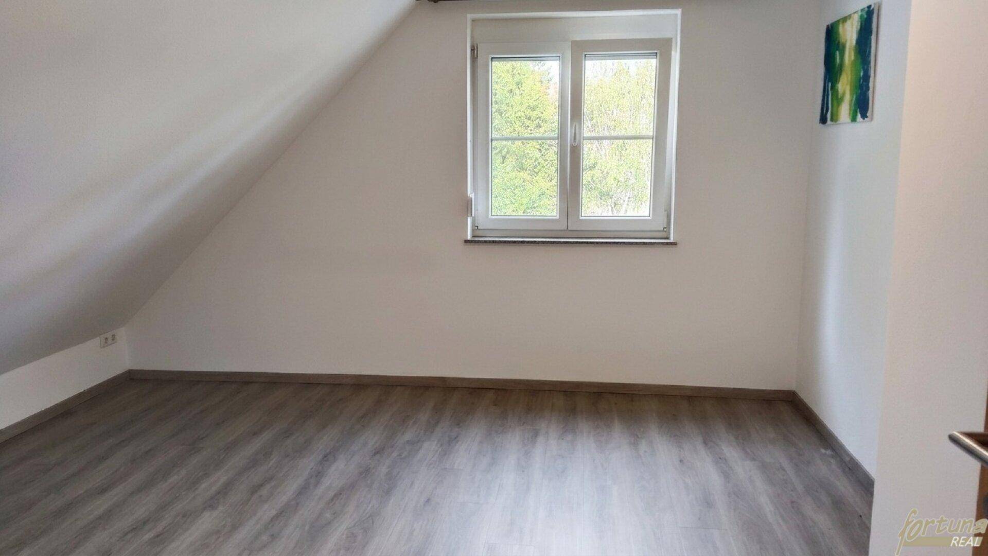 Leeres Zimmer im Dachgeschoss mit schräger Decke, Laminatboden und einem Fenster mit Blick ins Grüne.