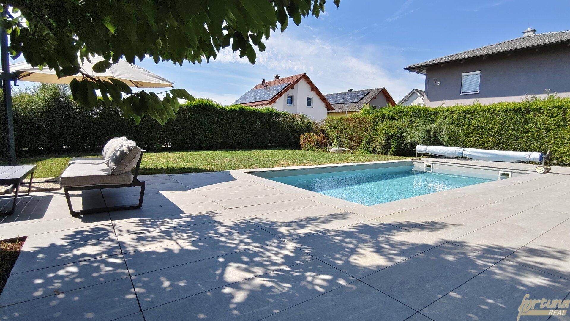 Gepflegter Gartenbereich mit Swimmingpool, gepflasterter Terrasse und Liegestühlen unter einem Sonnenschirm.