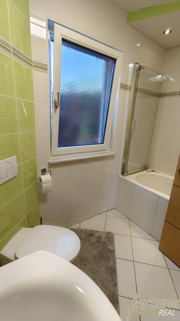Helles Badezimmer mit grünen Fliesen, Fenster und einer Badewanne mit Duschabtrennung.