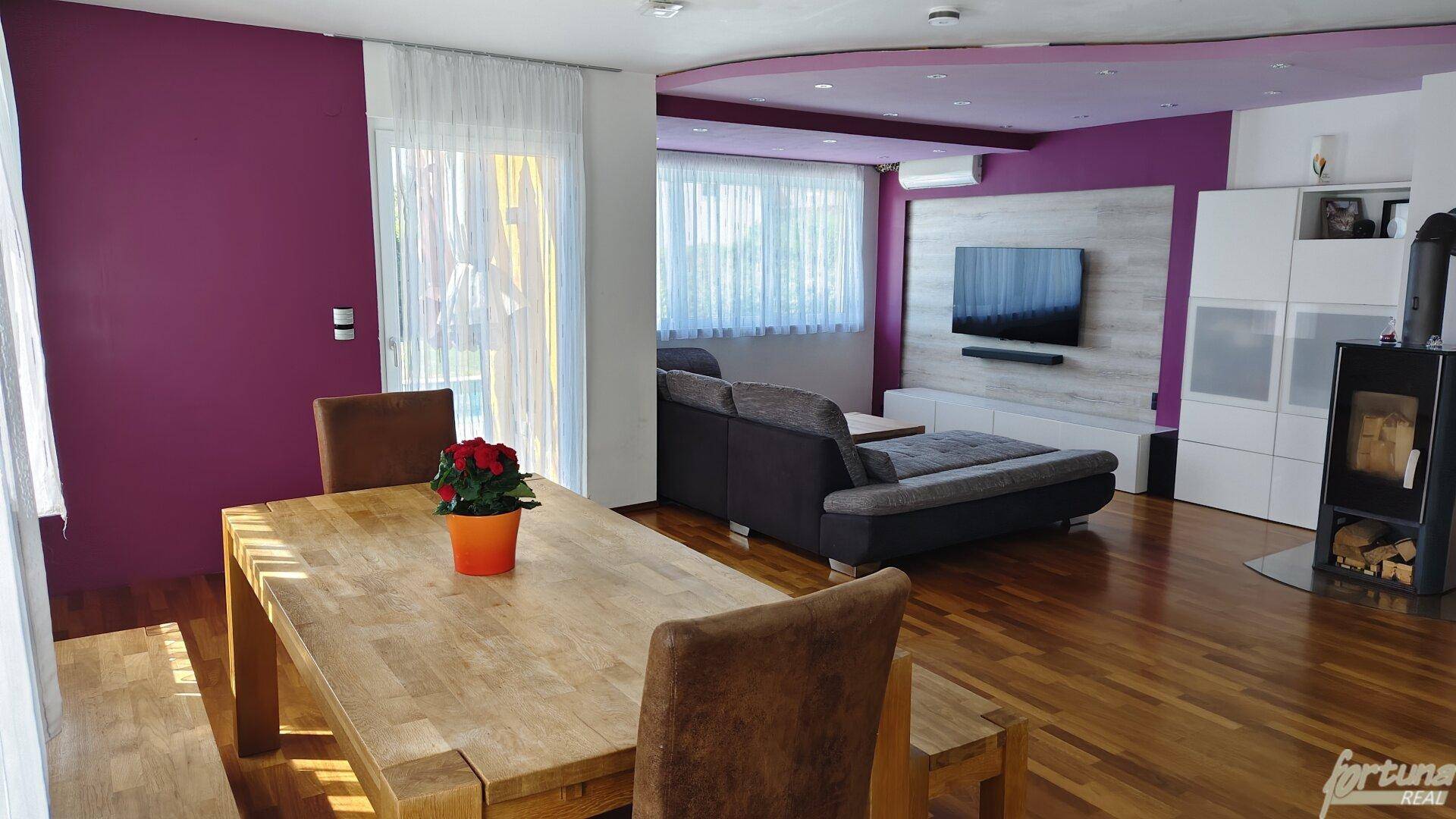 Heller Essbereich mit Holztisch und Blick auf den Wohnbereich mit farbigen Wänden und Fernseher.