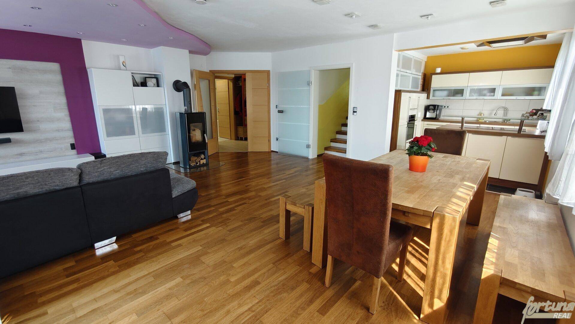 Offener Wohn-Essbereich mit Holzboden, Kaminofen und integrierter Küche im Hintergrund.