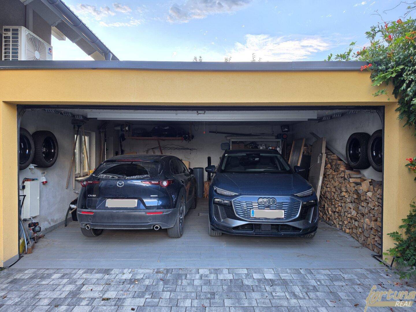 Geräumige Doppelgarage mit zwei Fahrzeugen, Holzstapel und zusätzlichen Reifenlagerflächen.