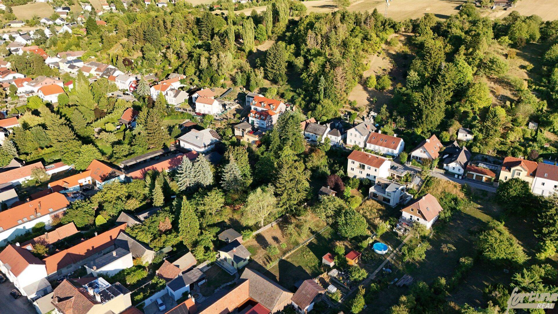Weitläufige Luftaufnahme einer ländlichen Siedlung, eingebettet in eine hügelige, grüne Landschaft mit vielen Häusern.