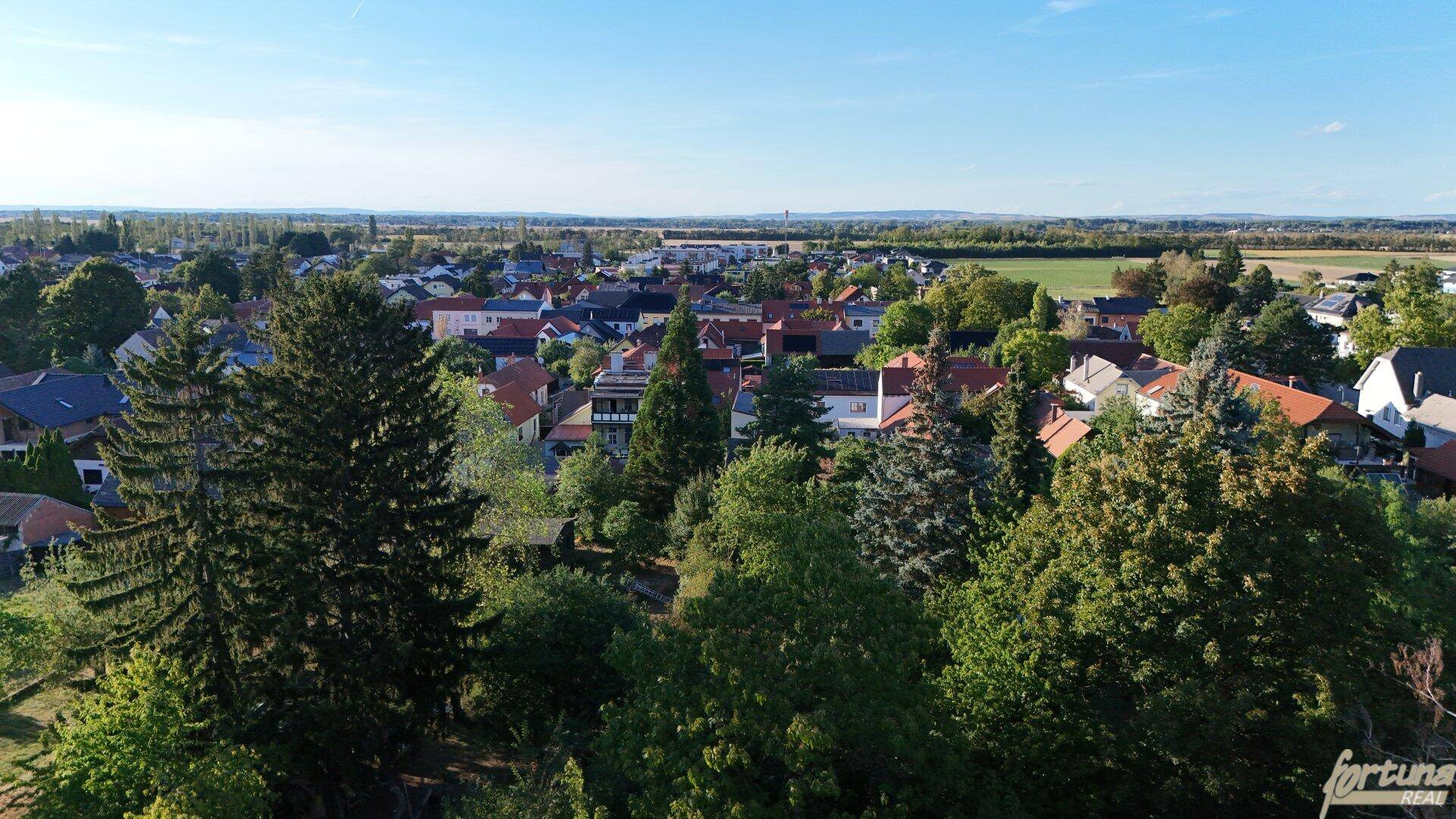 Luftaufnahme einer Wohnsiedlung, umgeben von üppigem Grün und weiten Feldern in der Ferne.