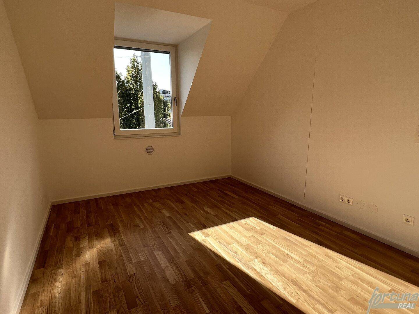 Helles Schlafzimmer mit Holzboden, Dachschräge und großem Fenster mit Blick ins Grüne.