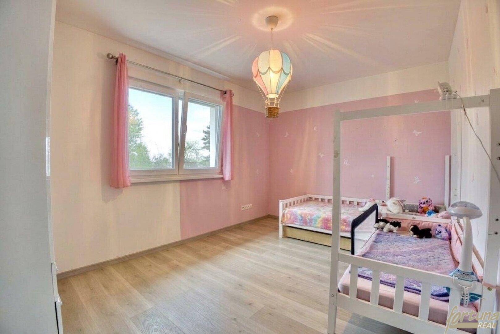 Kinderzimmer mit rosa Wänden, Laminatboden und zwei Einzelbetten unter einer bunten Deckenlampe.