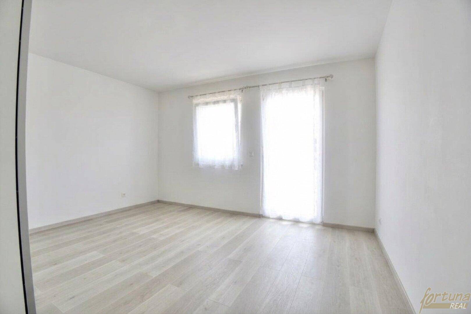 Leeres, helles Zimmer mit Laminatboden und großen Fenstern, bereit zur individuellen Gestaltung.