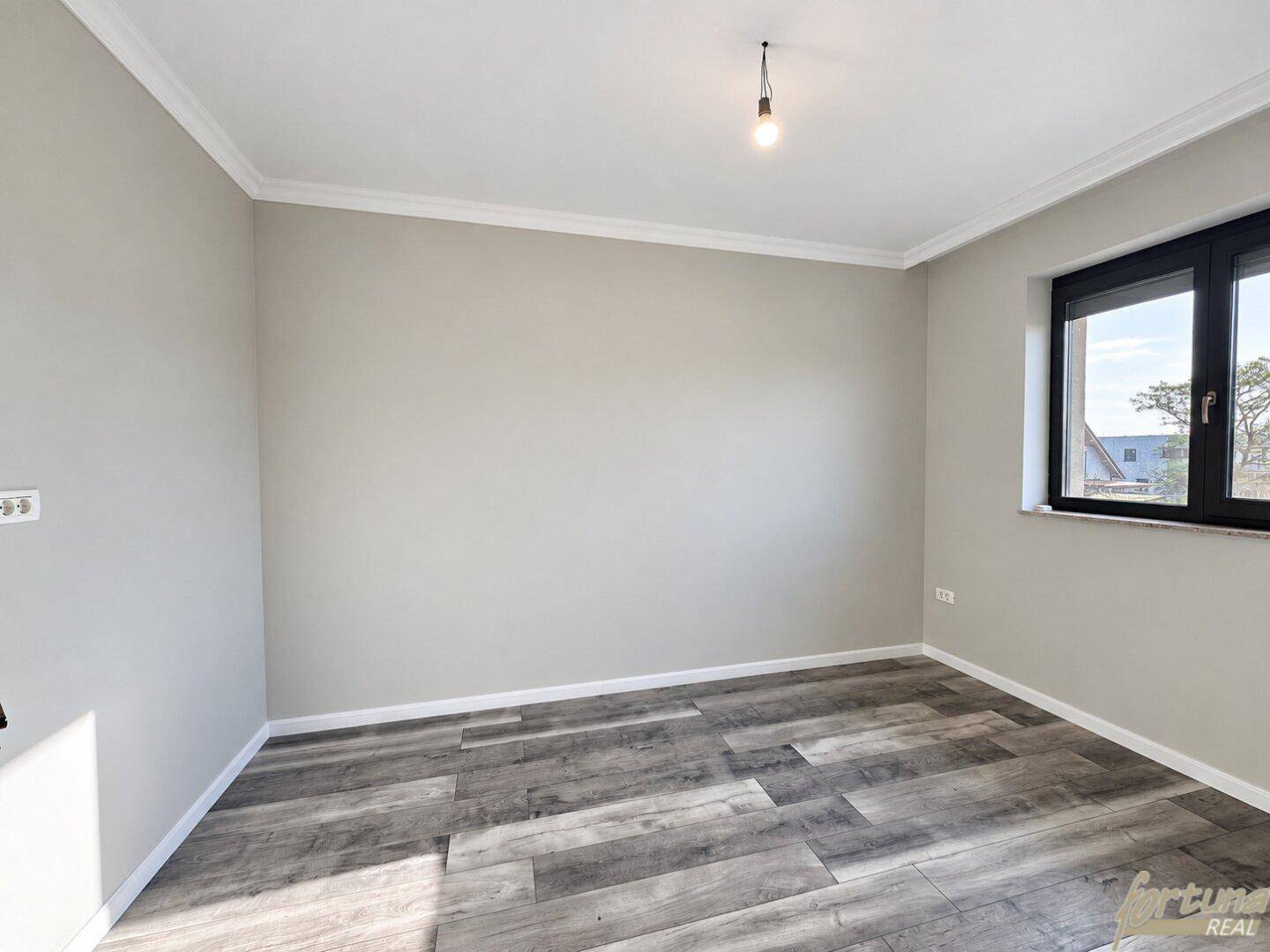 Helles, leeres Zimmer mit grauem Laminatboden und einem Fenster für natürliches Licht.
