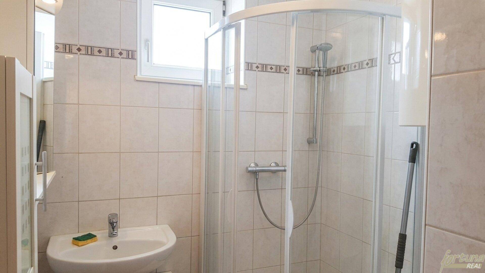 Badezimmer mit Duschkabine, Waschbecken und Fenster, komplett gefliest.