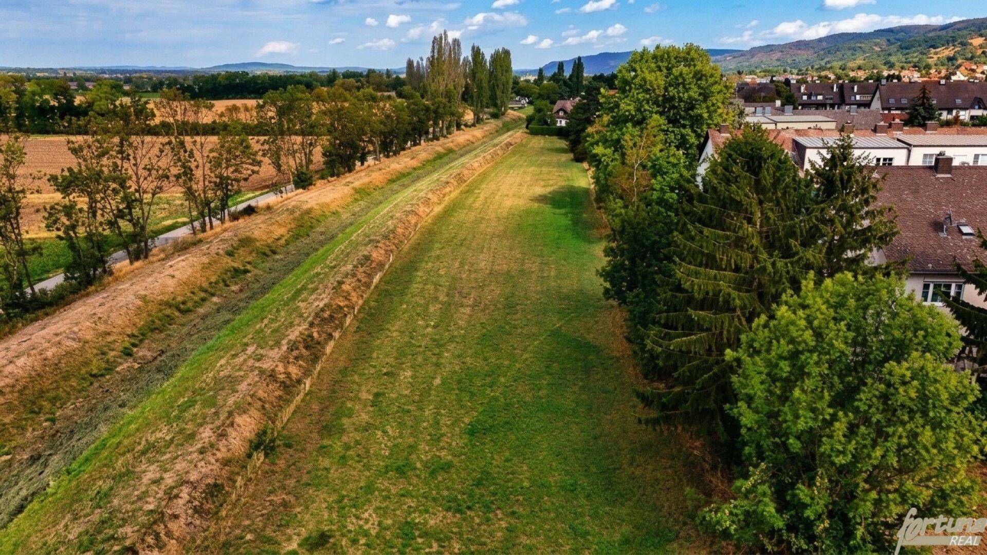 Weitläufige Grünfläche zwischen Feldern und einer Wohnbebauung unter blauem Himmel.