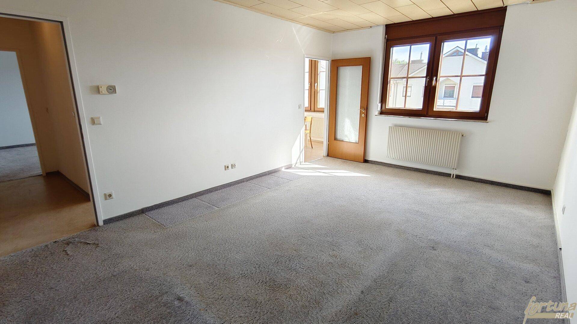Geräumiges Wohnzimmer mit Teppichboden, weißen Wänden und einem großen Fenster mit Heizkörper.