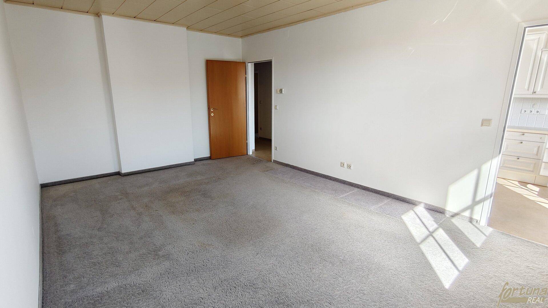 Geräumiger Raum mit Teppichboden und weißen Wänden, der zu einer angrenzenden Küche führt.