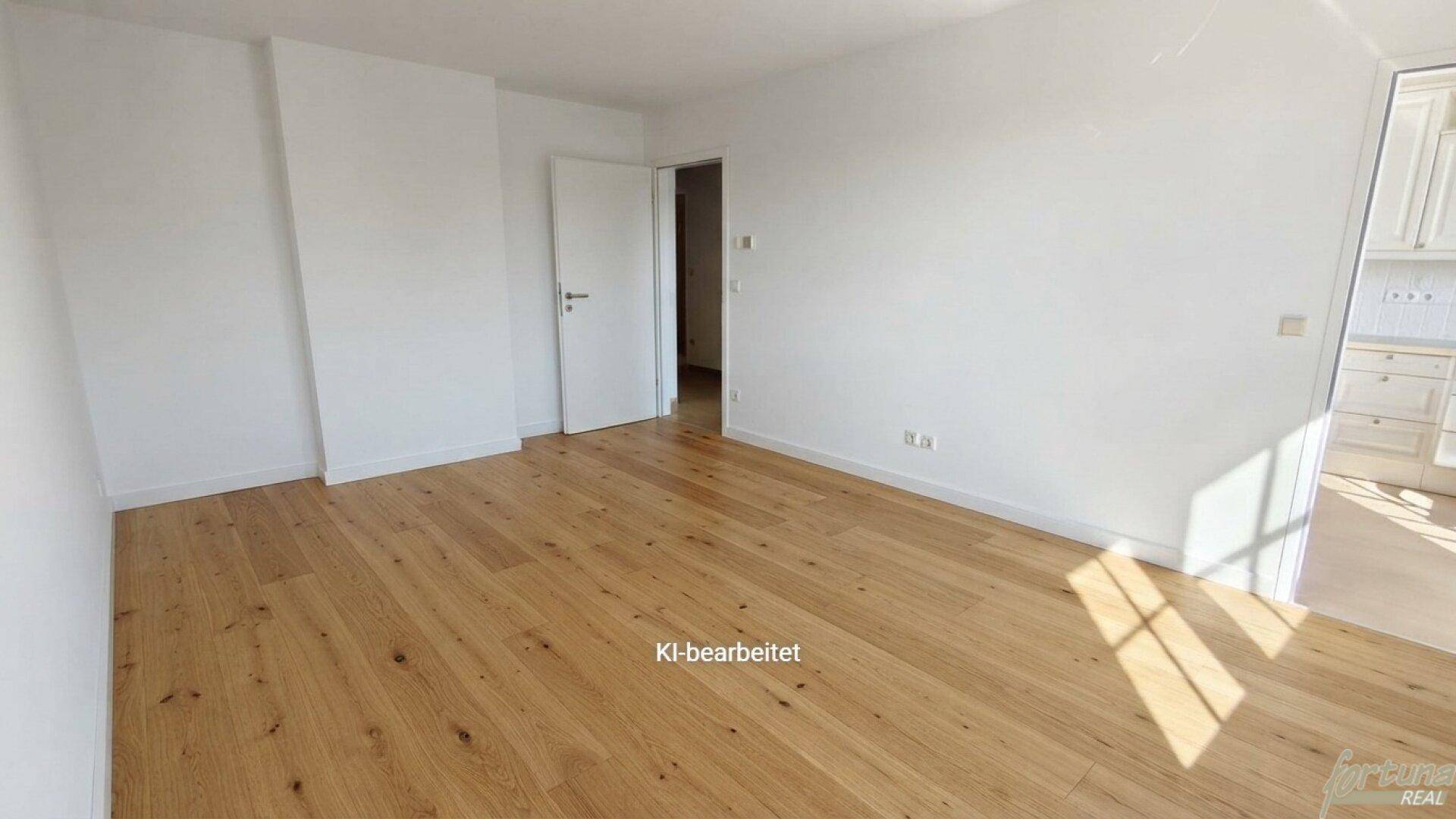 Heller Raum mit Holzboden und weißen Wänden, der zu einer angrenzenden Küche führt.