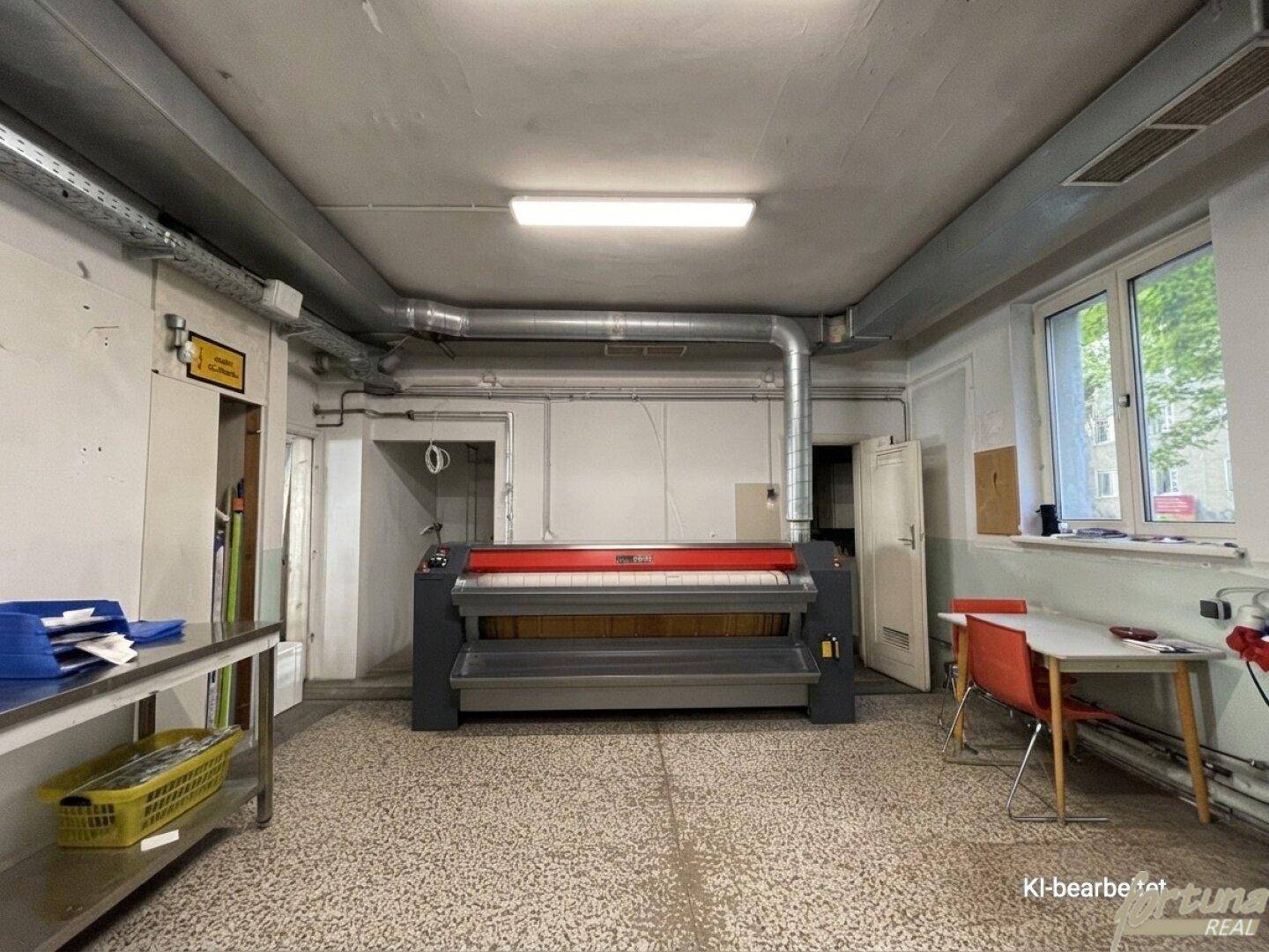 Industrielle Mangelmaschine im Zentrum eines großen Wäschereiraums, umgeben von Arbeitsflächen und Fenstern.