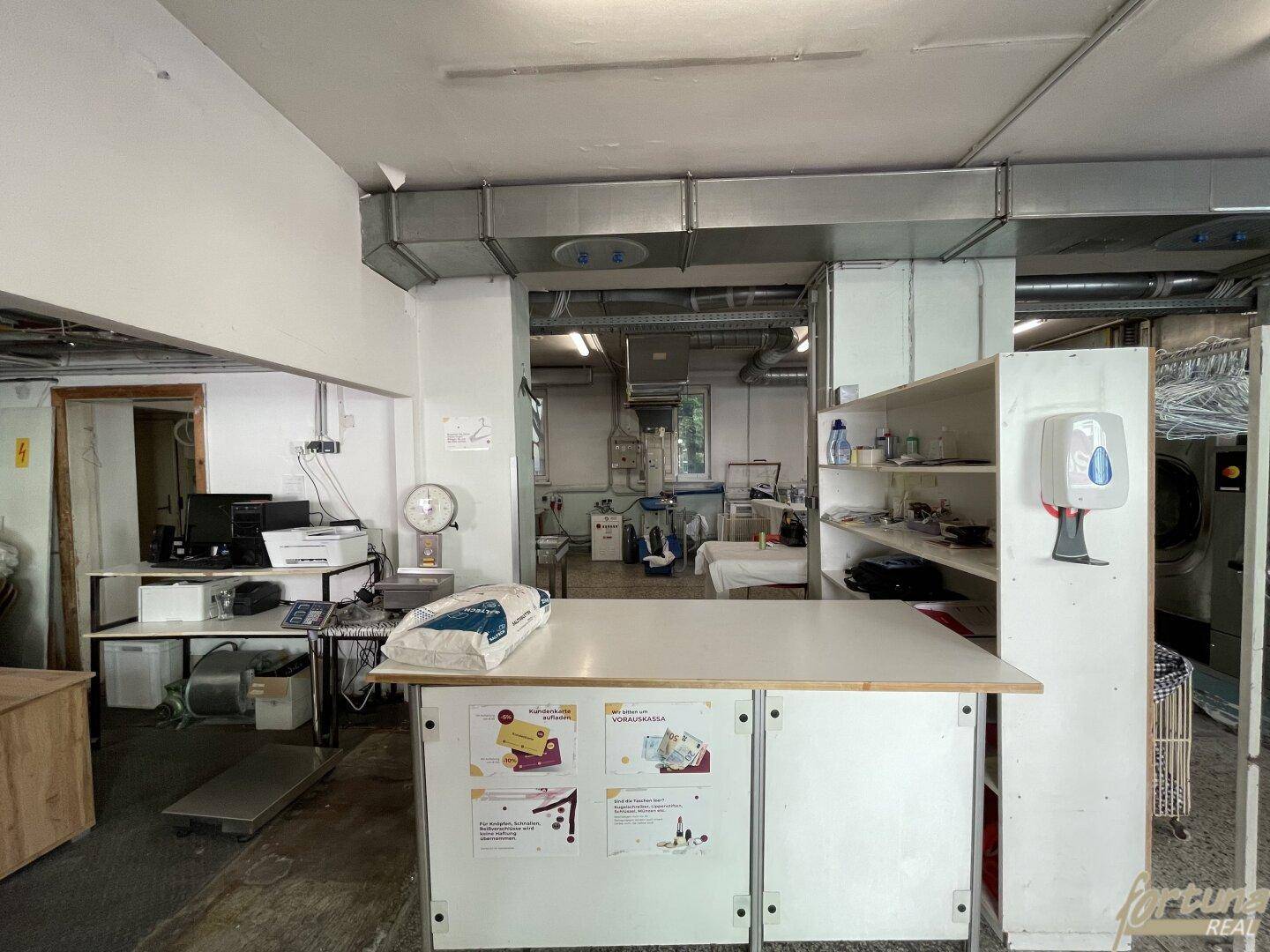 Empfangsbereich oder Kasse in der Wäscherei mit Schreibtisch, Computer und Regalen, mit Blick auf den Arbeitsbereich.