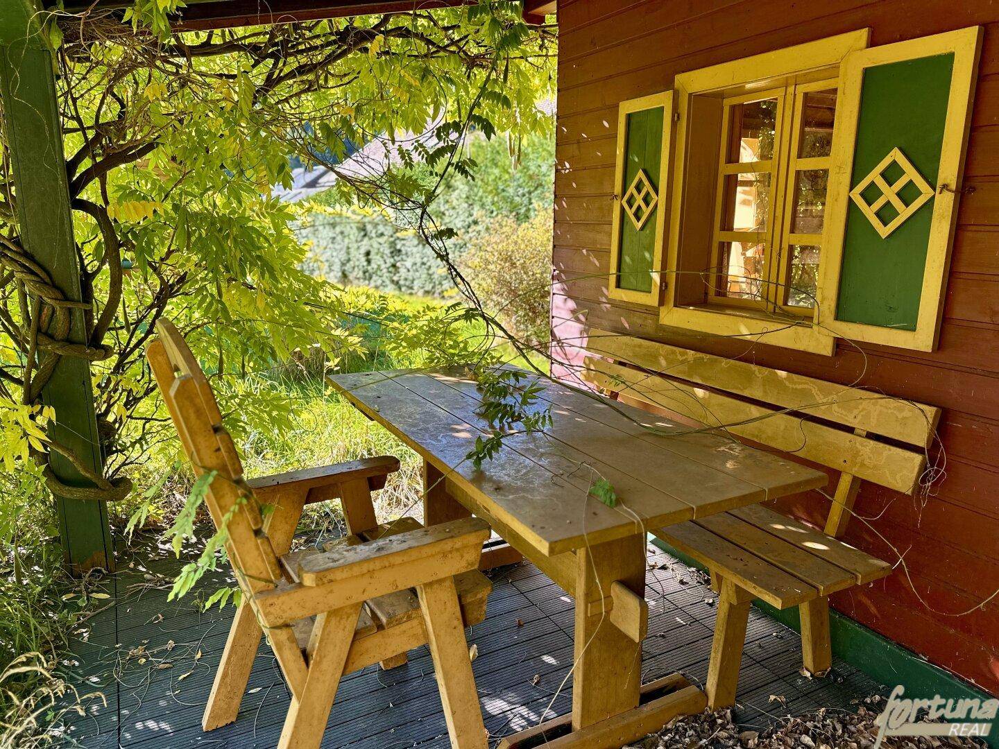 Gemütliche Terrasse mit rustikalen Holzmöbeln und grünen Fensterläden, umgeben von üppigem Grün.
