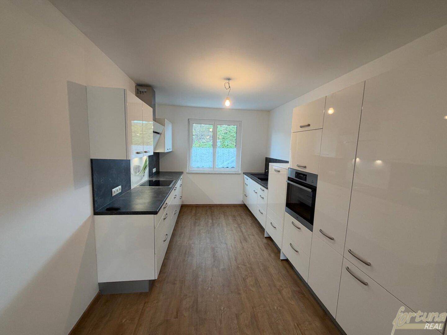 Moderne Küche mit weißen Hochglanzfronten, dunklen Arbeitsplatten und Fensterblick ins Grüne.