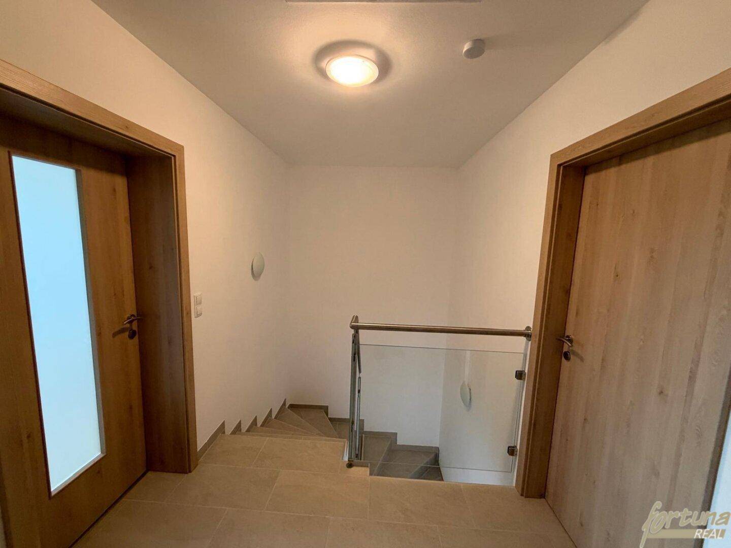 Heller Treppenaufgang mit modernem Glasgeländer und Fliesenboden im Obergeschoss.