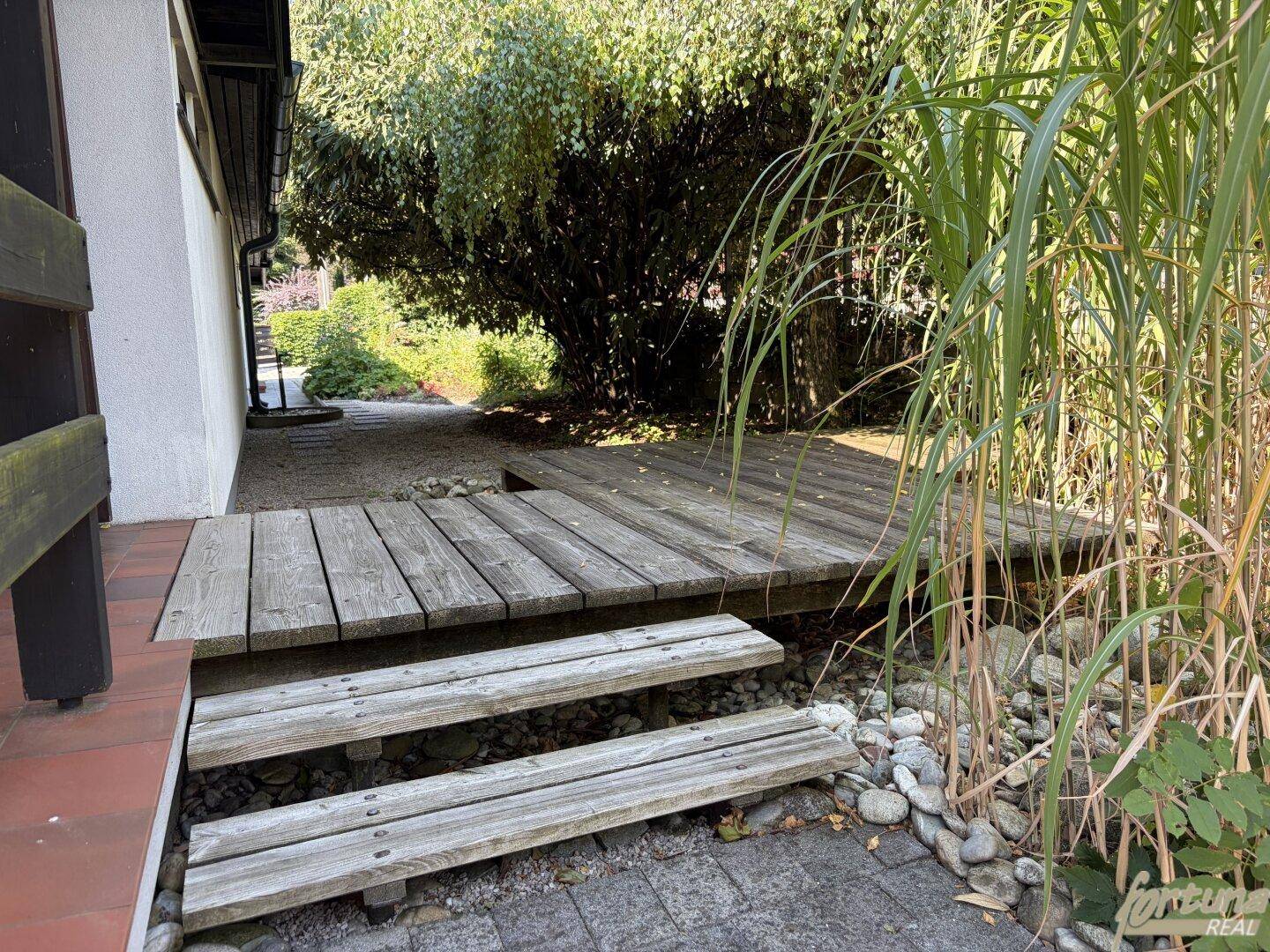 Gartenbereich mit Holzstegen und Kieswegen, umgeben von üppigem Grün und Bäumen.