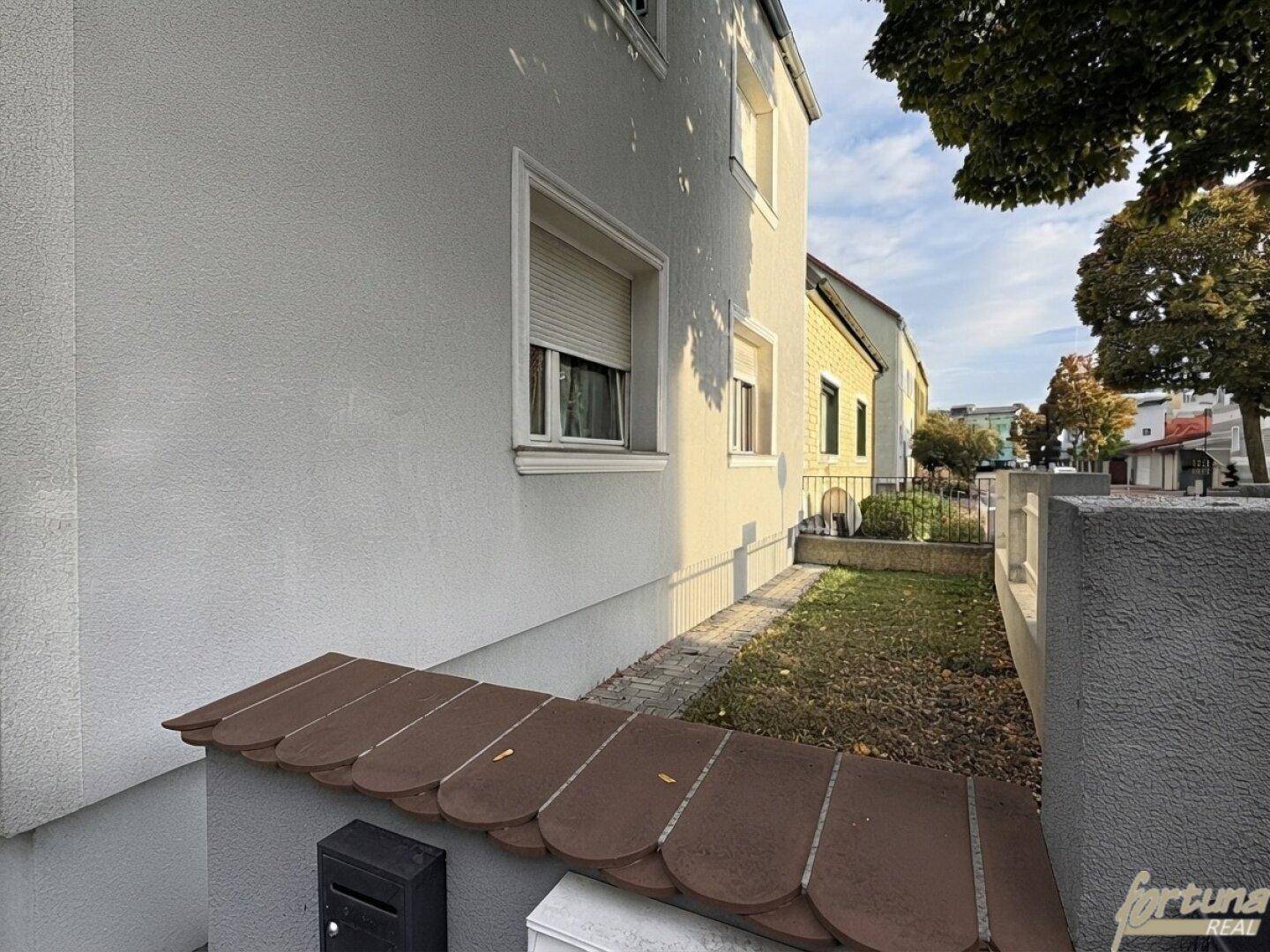 Seitenansicht des Gebäudes mit Fenstern, Rollläden und einem kleinen Gartenbereich zur Straße hin.
