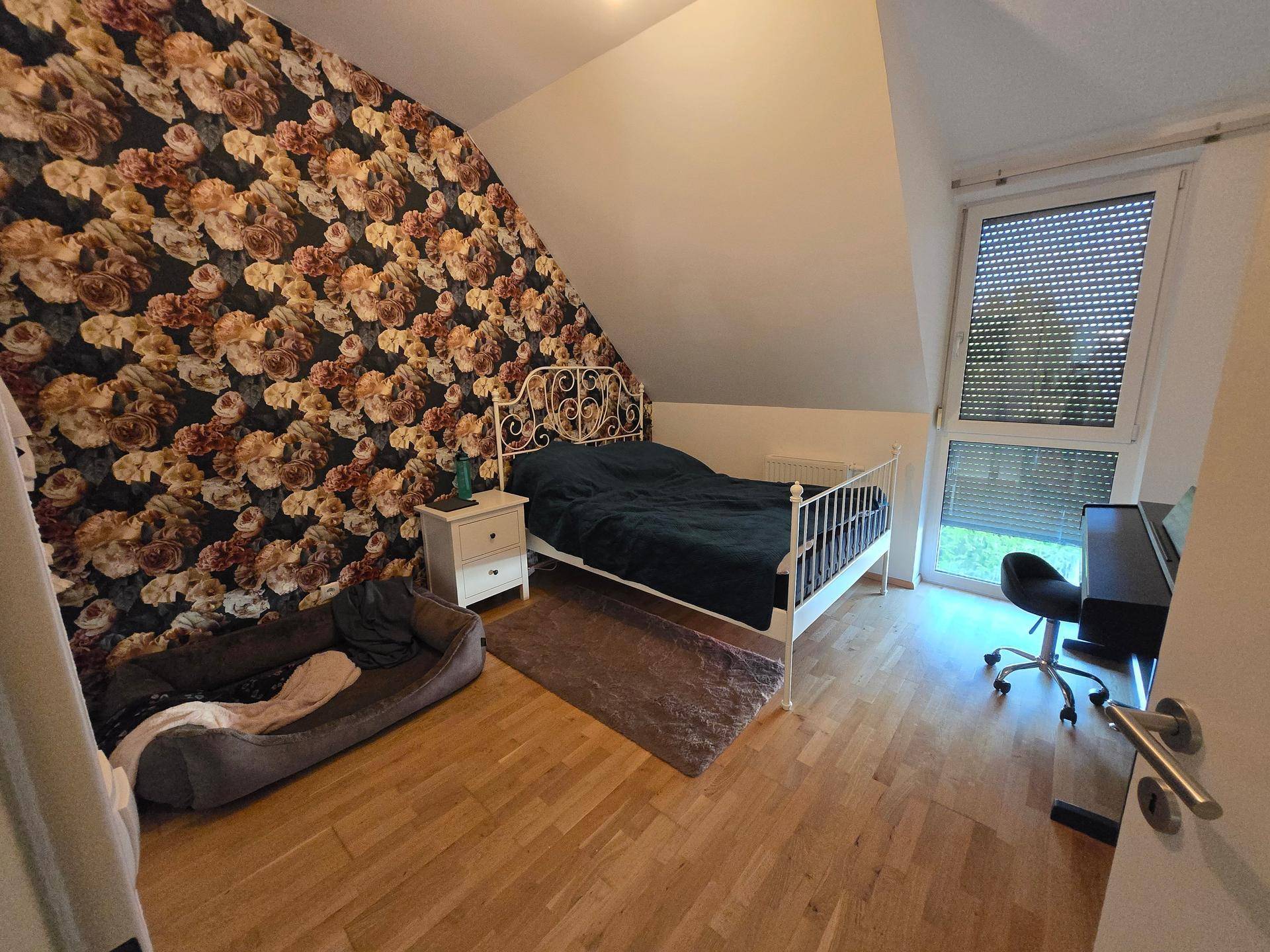 Schlafzimmer mit auffälliger Blumentapete, Holzboden und großem Fenster für viel Licht.