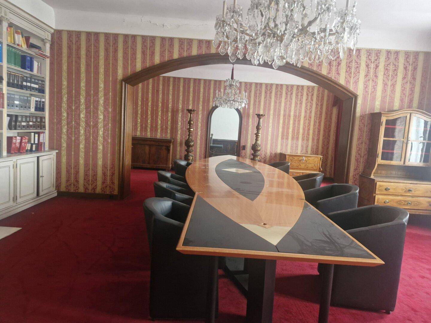 Großzügiges Wohnzimmer mit rotem Teppichboden, Bücherregalen und einem dekorativen Kamin, beleuchtet von einem Kronleuchter.