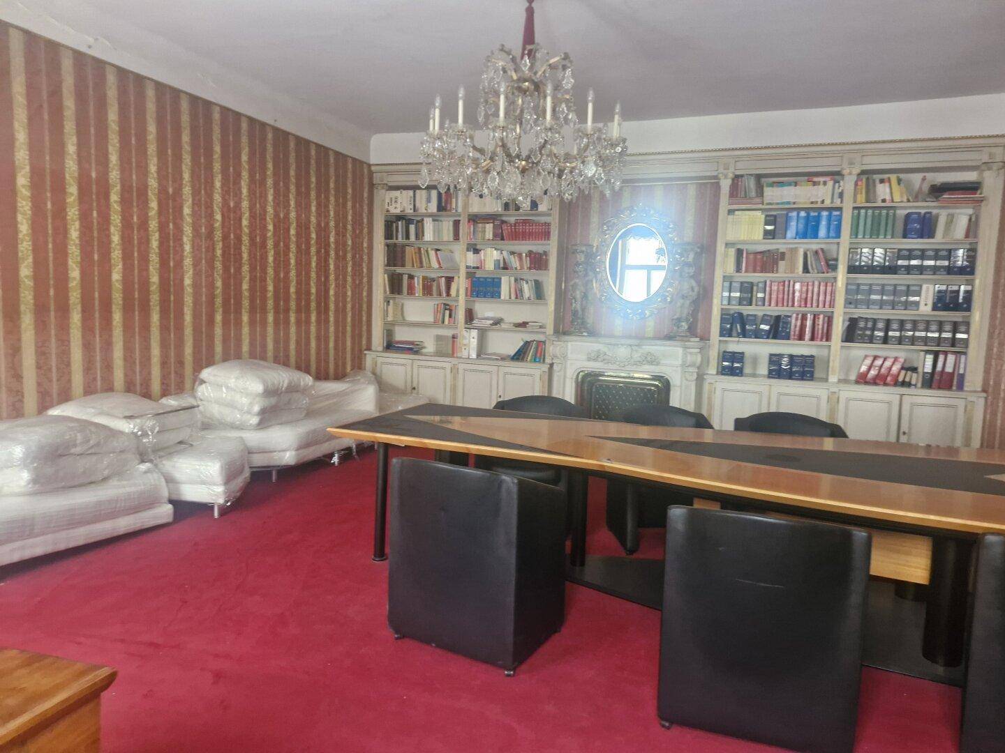 Wohnzimmer mit rotem Teppichboden, Kronleuchter, Bücherregalen und einem großen Tisch mit Stühlen.