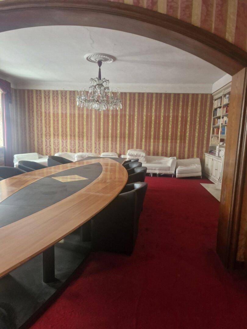 Blick durch einen Bogen in ein Wohnzimmer mit rotem Teppichboden und einem eleganten Kronleuchter.