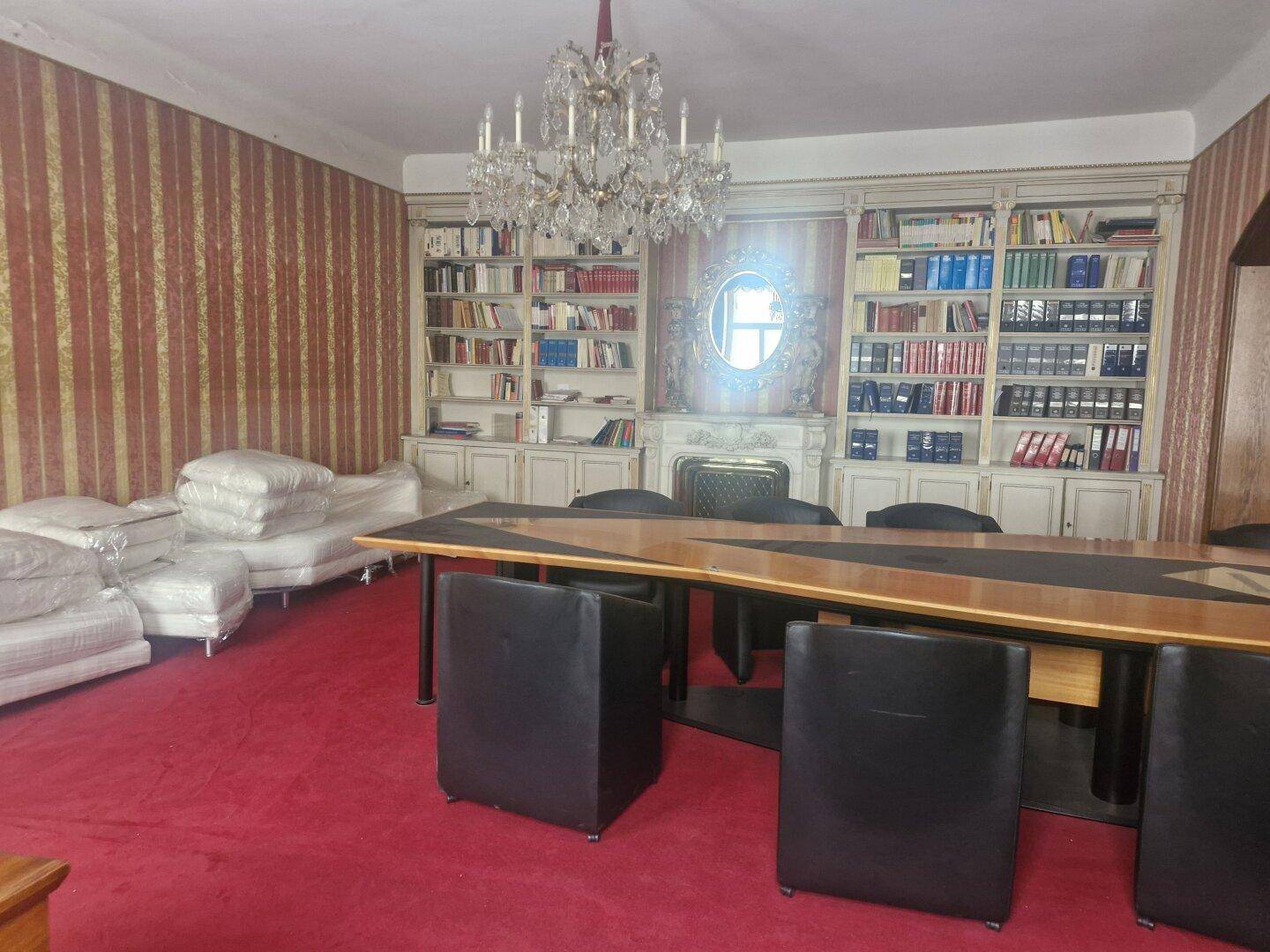 Großzügiges Wohnzimmer mit rotem Teppichboden, Bücherregalen und einem dekorativen Kamin, beleuchtet von einem Kronleuchter.