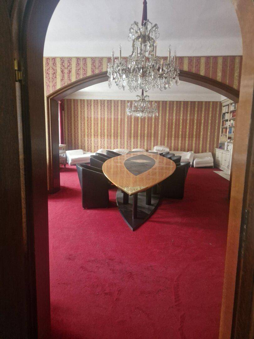 Elegantes Wohnzimmer mit rotem Teppichboden, Kronleuchter und einem modernen Tisch in der Mitte des Raumes.