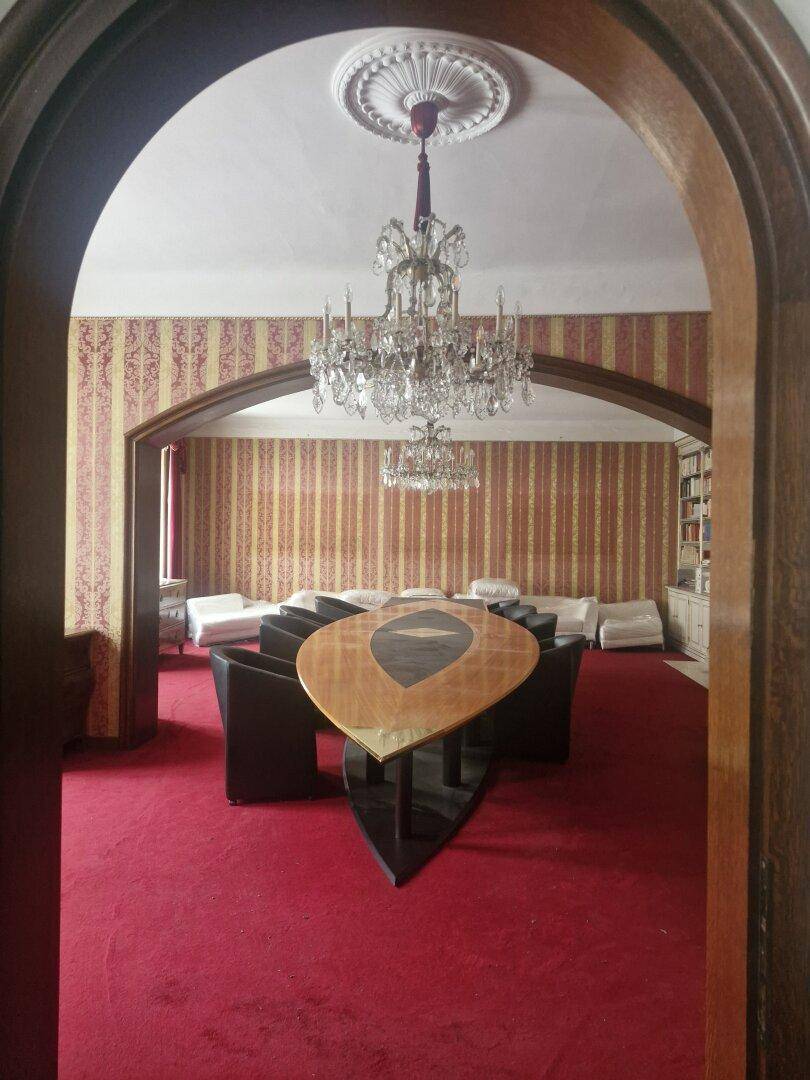 Elegantes Wohnzimmer mit rotem Teppichboden, Kronleuchter und einem modernen Tisch in der Mitte des Raumes.
