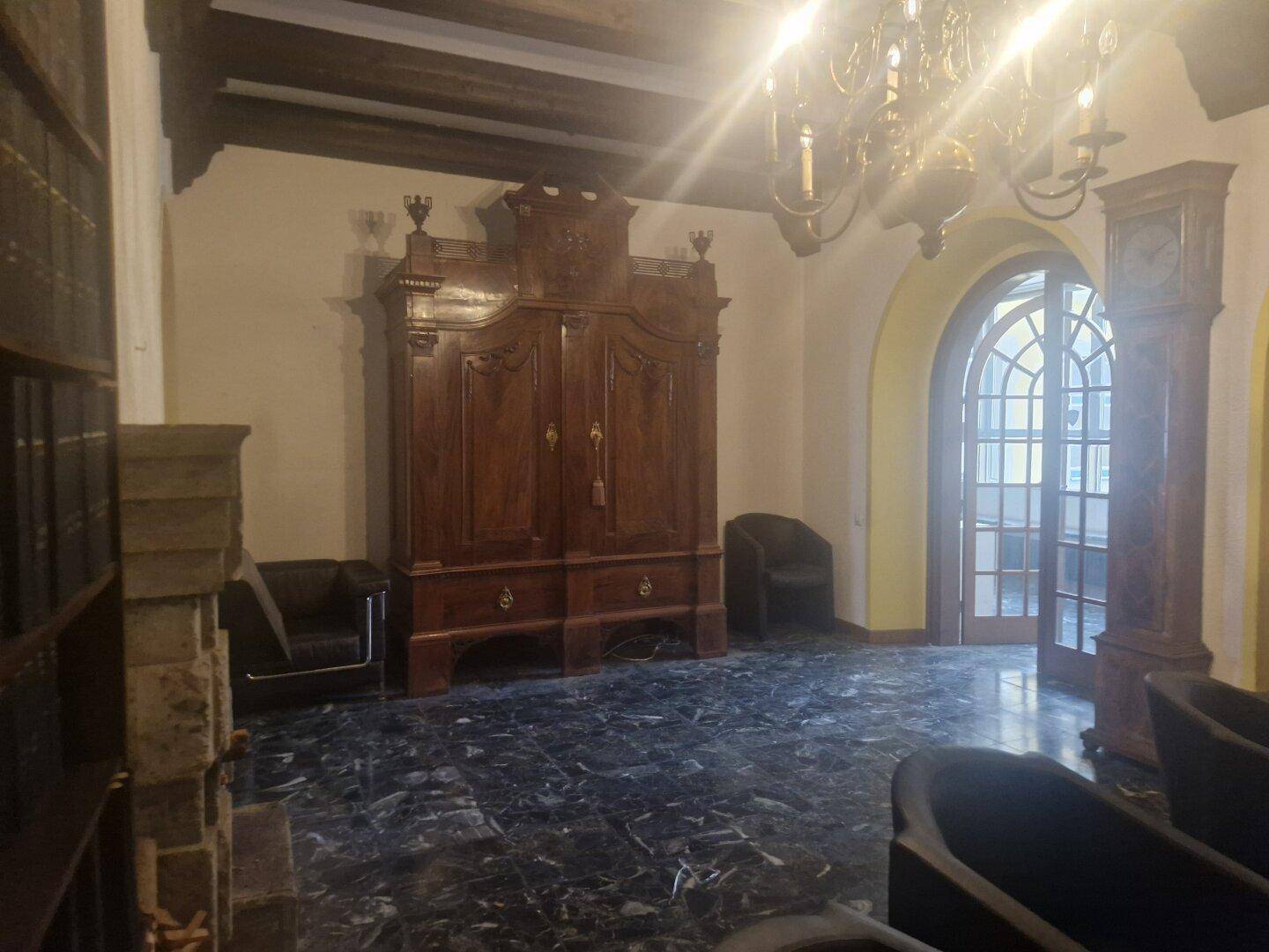 Wohnzimmer mit dunklem Marmorboden, einem großen Holzschrank und einem Kronleuchter, umgeben von Sesseln.
