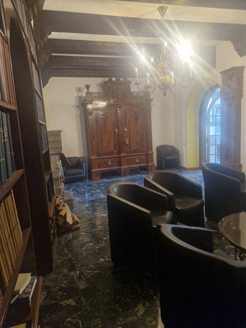 Wohnzimmer mit dunklem Marmorboden, einem großen Holzschrank und einem Kronleuchter, umgeben von Sesseln.