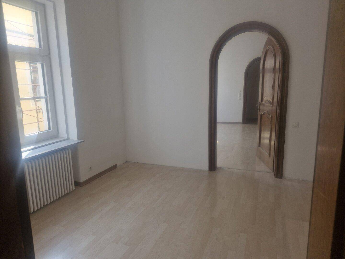 Leerer Raum mit hellem Boden, weißen Wänden und einem Fenster mit Heizkörper, Blick auf Rundbogen-Türen.