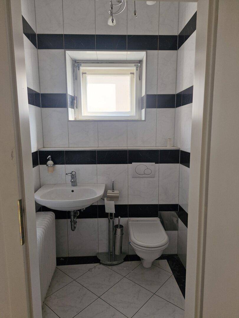 Kleines Gäste-WC mit Fenster, weiß-schwarz gefliesten Wänden und Boden, sowie einem Waschbecken und WC.