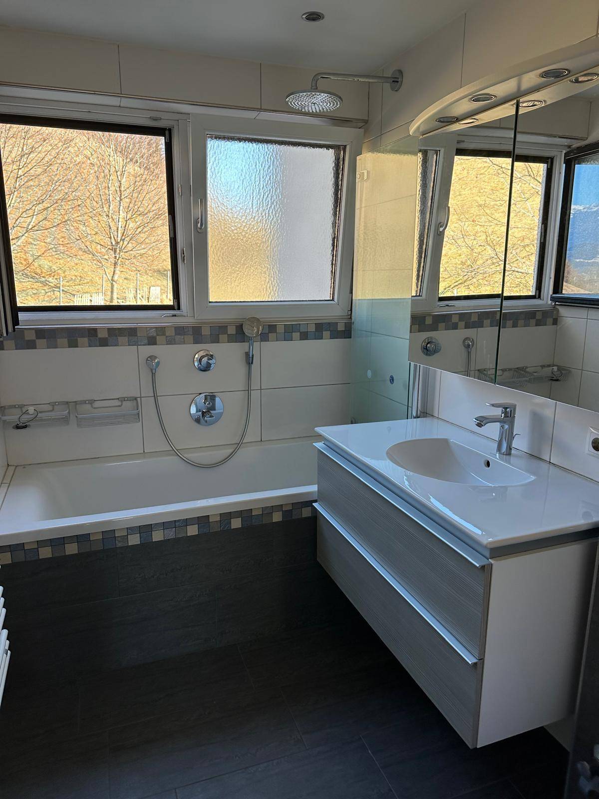 Helles Badezimmer mit Badewanne, Dusche, modernem Waschtisch und zwei Fenstern.