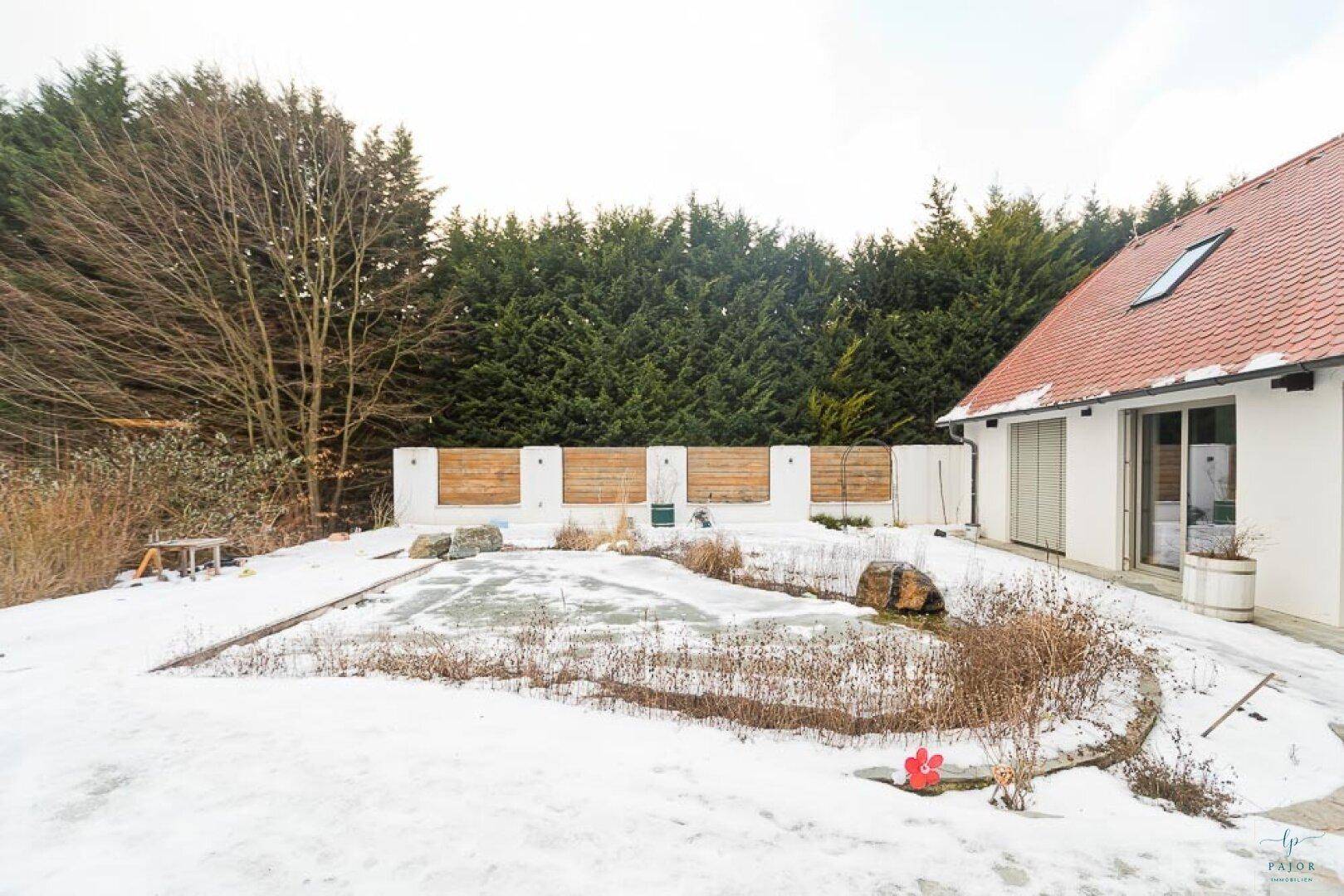 Weitläufiger winterlicher Garten mit Teich, umgeben von Bäumen und einer modernen Zaunanlage.
