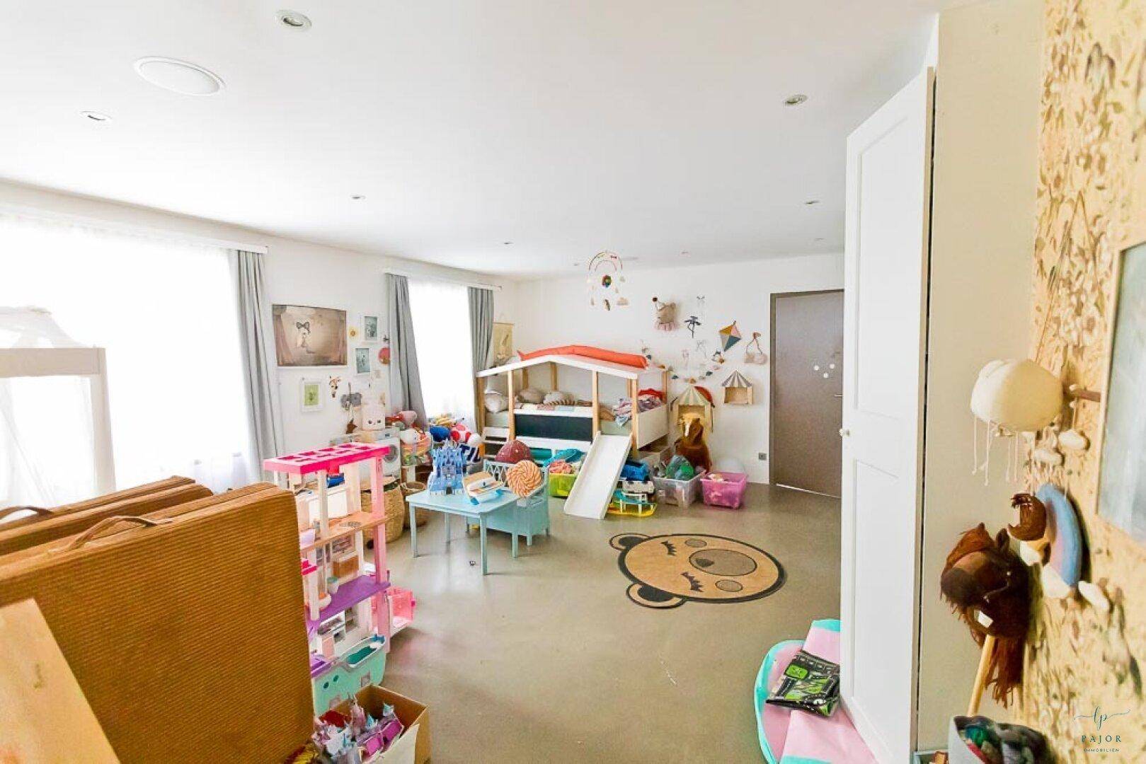 Großes, helles Kinderzimmer mit vielen Spielsachen, einem kleinen Bett und großen Fenstern.