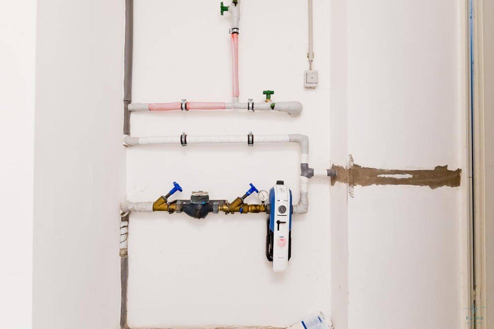 Installationsbereich mit sichtbaren Wasserleitungen und Zählern an einer weißen Wand.