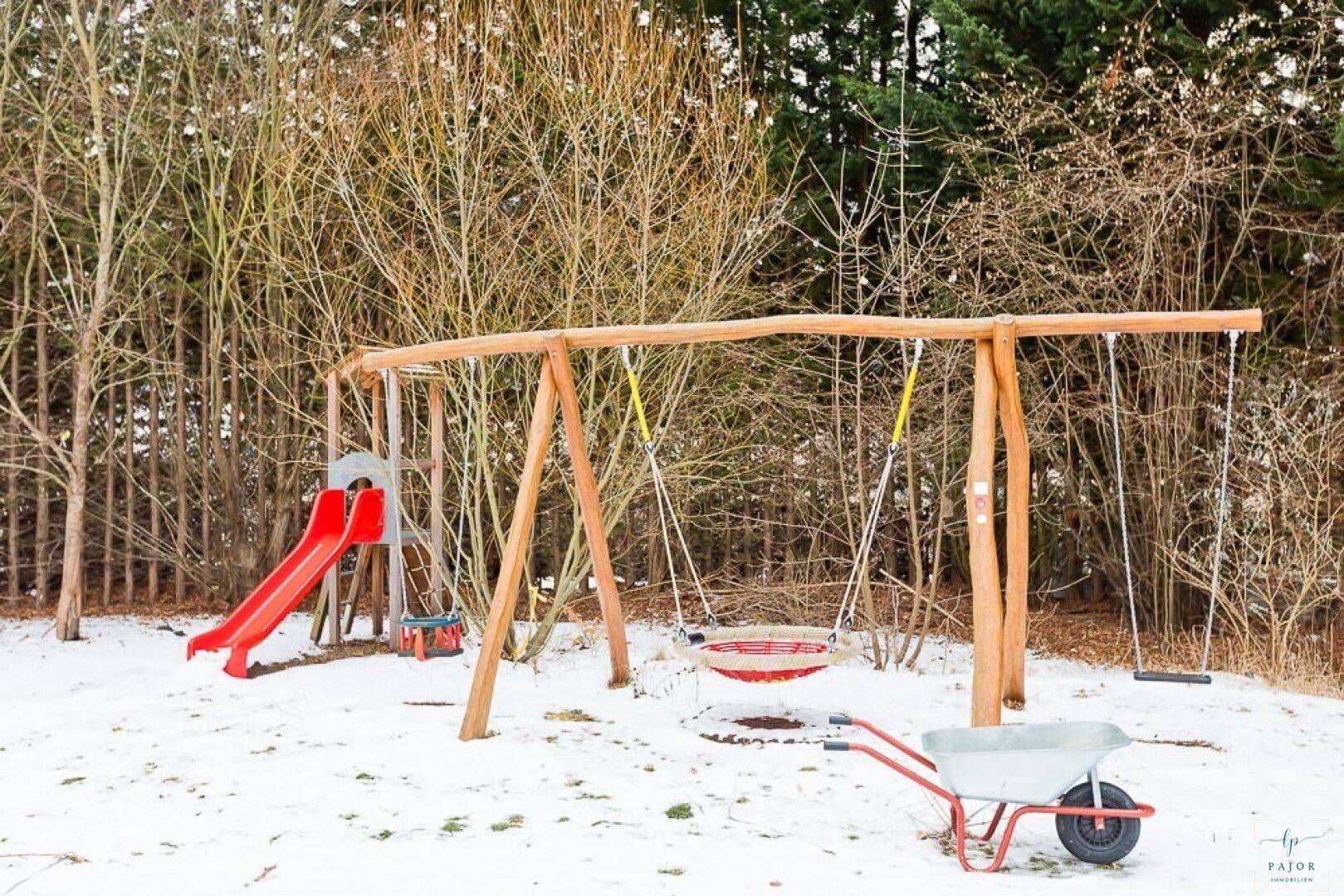 Winterlicher Garten mit einem Holzspielplatz, Rutsche und Schaukeln im Schnee.