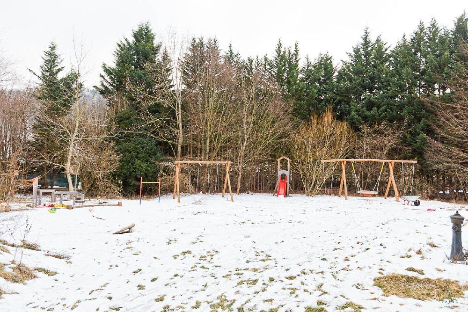 Weitläufiger Gartenbereich mit mehreren Spielgeräten und Bäumen, bedeckt mit Schnee.