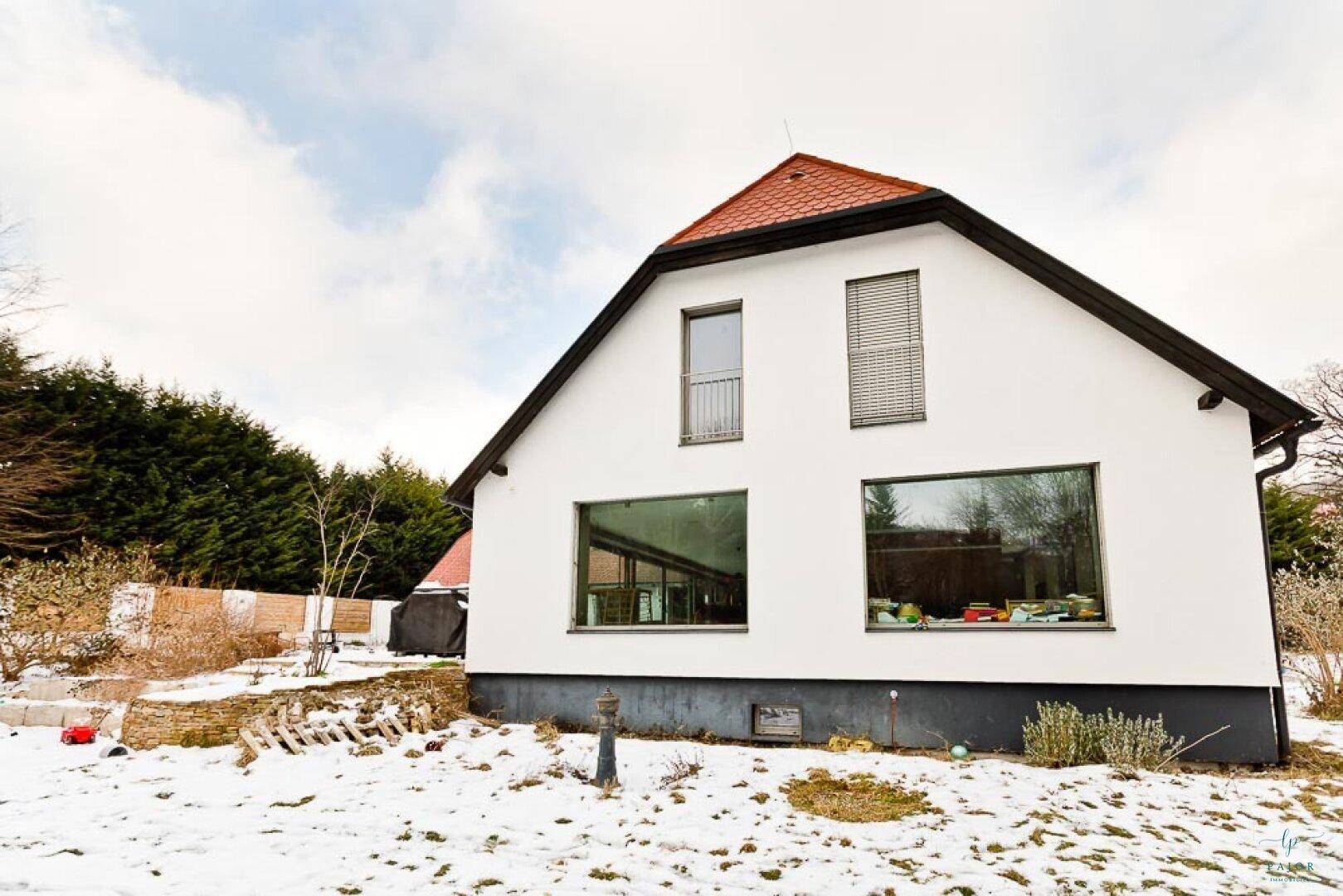 Modernes weißes Haus mit großen Fenstern und rotem Dach, umgeben von Schnee.