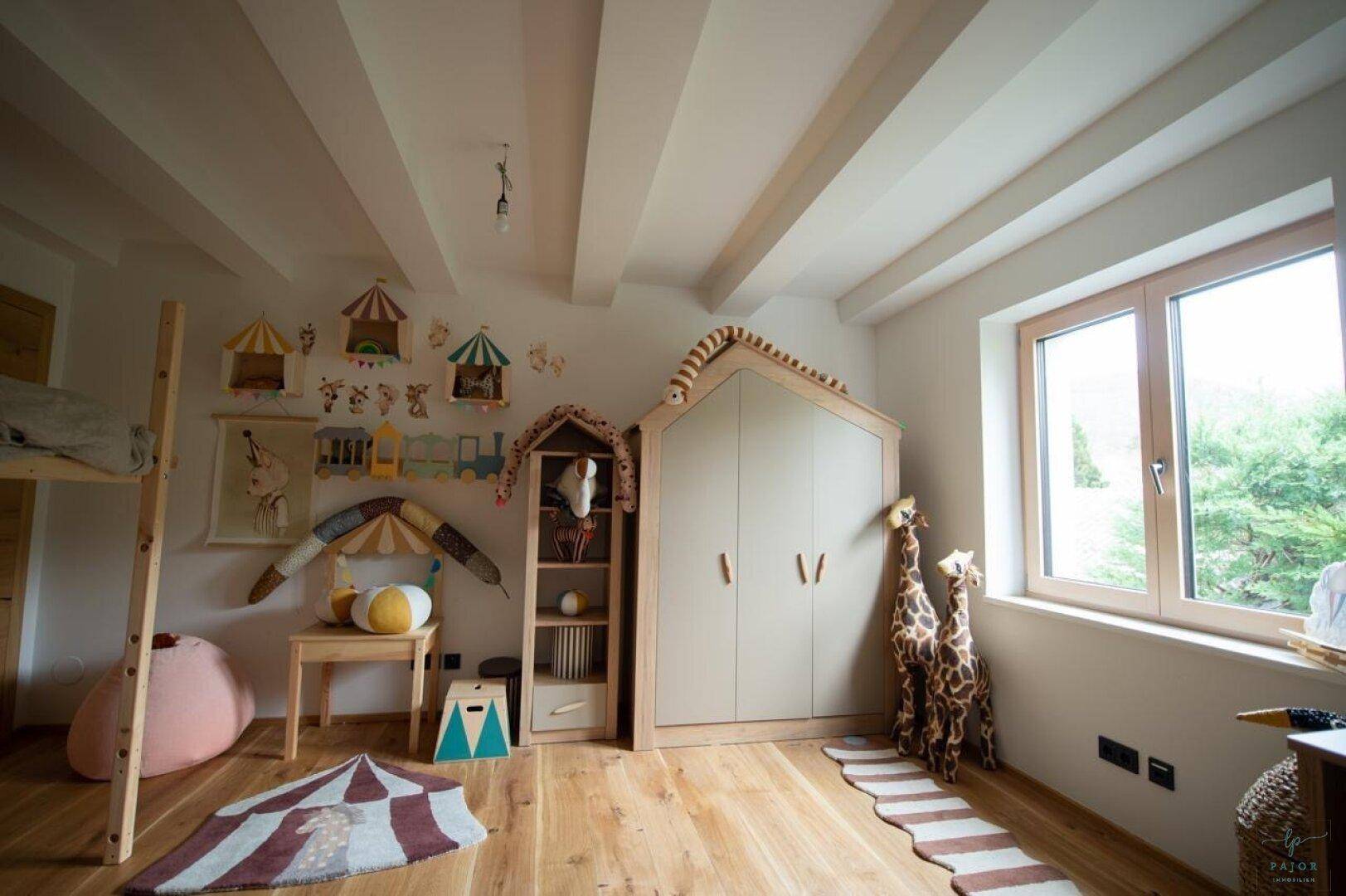 Helles Kinderzimmer mit Holzboden, sichtbaren Deckenbalken und einem großen Fenster.